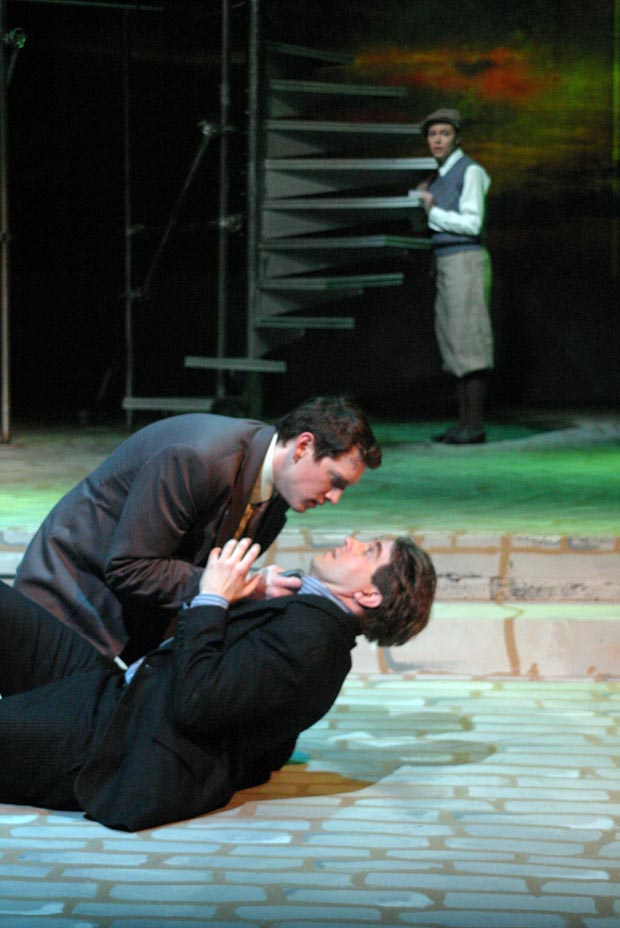 Intensity on stage: A man kneels, gripping another by the collar, his expression fierce, while a distant figure observes the confrontation.