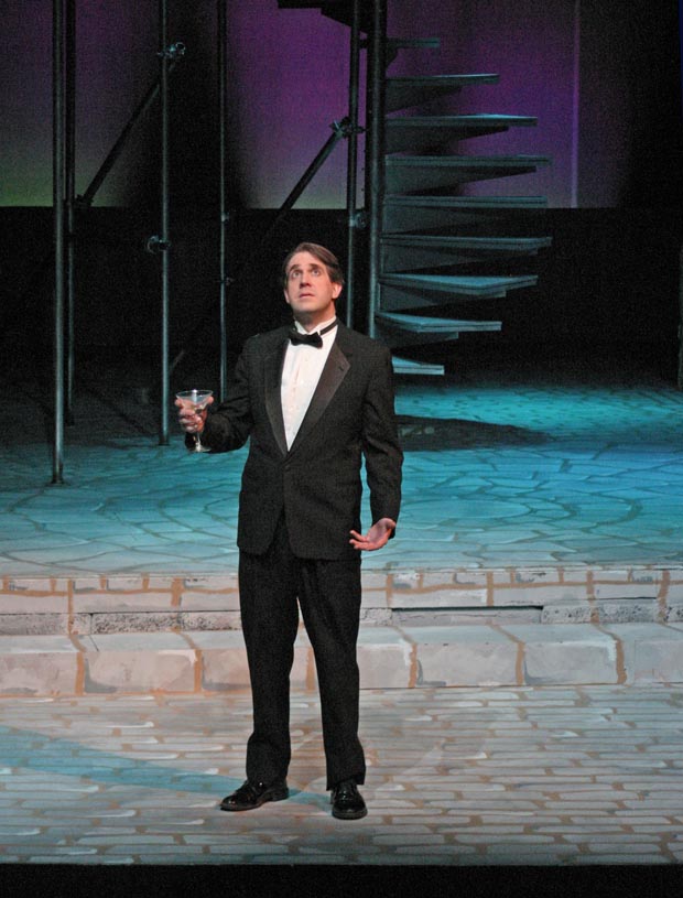 On stage, a man dressed in a tuxedo stands, holding a glass of champagne and looking upward with a contemplative expression.