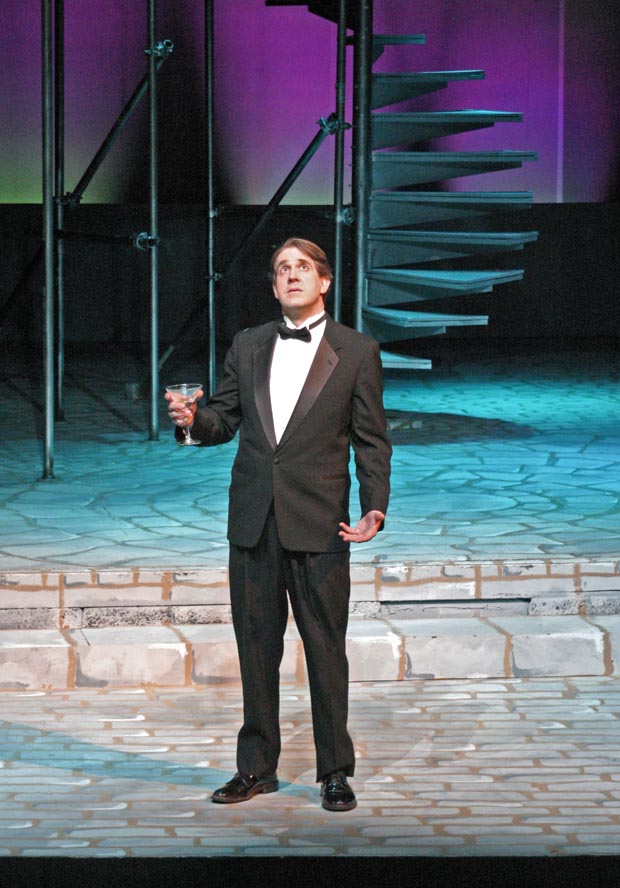 On stage, a man dressed in a tuxedo stands, holding a glass of champagne and looking upward with a contemplative expression.