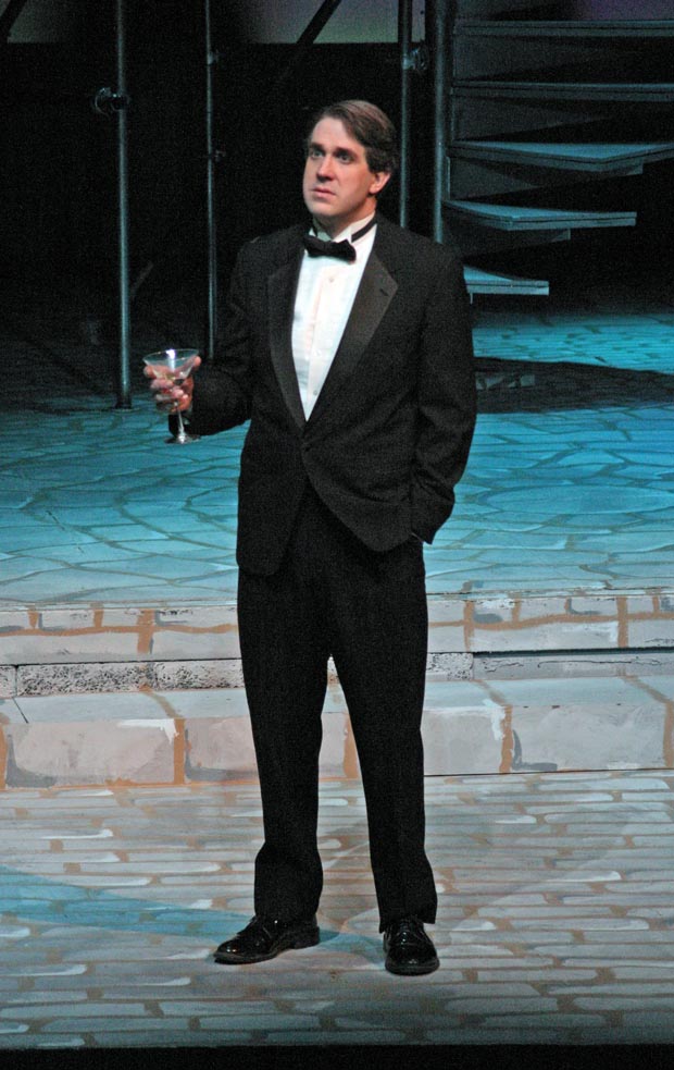 On stage, a man dressed in a tuxedo stands, holding a glass of champagne and looking towards audience with a contemplative expression.