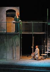 On stage, a woman dressed in green on a balcony looking down upon a man dressed in a suit sitting on the stairs.
