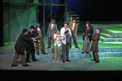 Trapped in the spotlight's glare, a terrified couple confronts a menacing semi-circle of seven men, some brandishing weapons, as uncertainty hangs heavy in the air on stage.