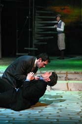 Intensity on stage: A man kneels, gripping another by the collar, his expression fierce, while a distant figure observes the confrontation.