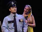 Two actors on stage during the Winter Shorts production of Cop-Out, one dressed as a police officer and another as a woman who is standing behind him, smiling with her hands on his shoulders | UAF Photo by Kade Mendelowitz