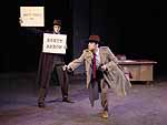 Two actors on stage during the Winter Shorts production of Cop-Out, one os a man in a suit holding two signs that say "Matt Krell as Brett Arrow." The other man is in a trenchcoat with his arm extended, holding a gun, mid run across the stage | UAF Photo by Kade Mendelowitz