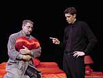 Actors on stage during the Winter Shorts performance of Panic in the Time of an Insecure God. Two men are talking. One is sitting on a bed holding a heart pillow. The other is standing and pointing at him | UAF Photo by Kade Mendelowitz