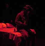 Actor on stage during the Winter Shorts performance of Panic in the Time of an Insecure God. Dramatic red light shines down on a man sitting on a bed wearing a trenchcoat | UAF Photo by Kade Mendelowitz