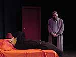 Actors on stage during the Winter Shorts performance of Panic in the Time of an Insecure God. A man lays back on a bed in exhaustion with his hands on his heads, while a man in a robe smiles down at him | UAF Photo by Kade Mendelowitz