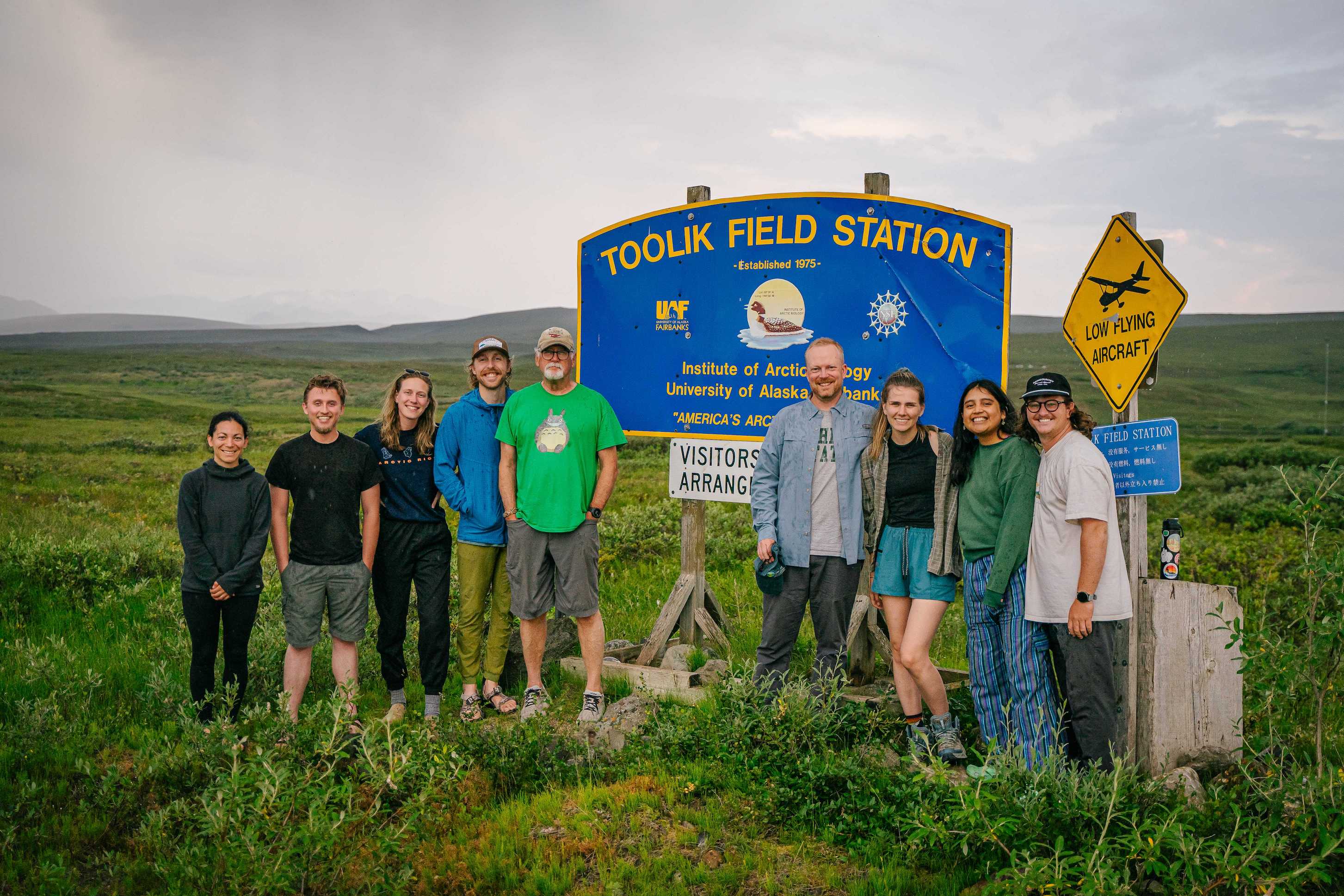 Scientists stand by the Toolik Field Station sign on a sunny, summer day