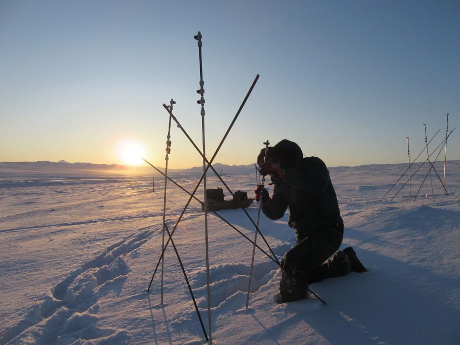 A woman crouches near three permafrost gas sampling poles bored into the snow covered tundra on a sunny November day.