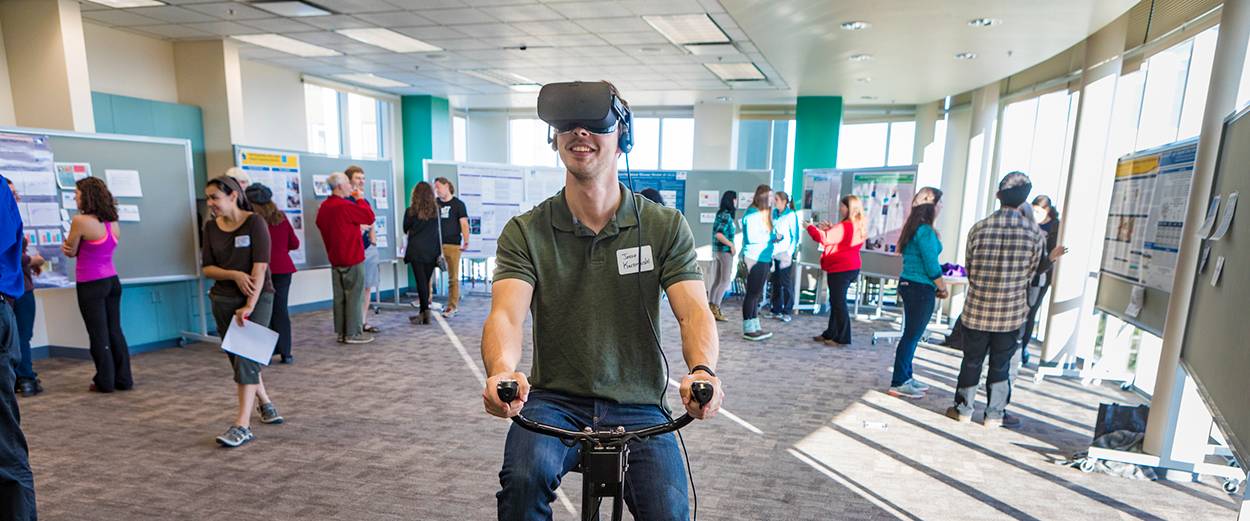 A UAF student on a stationary bike wearing a VR headset.