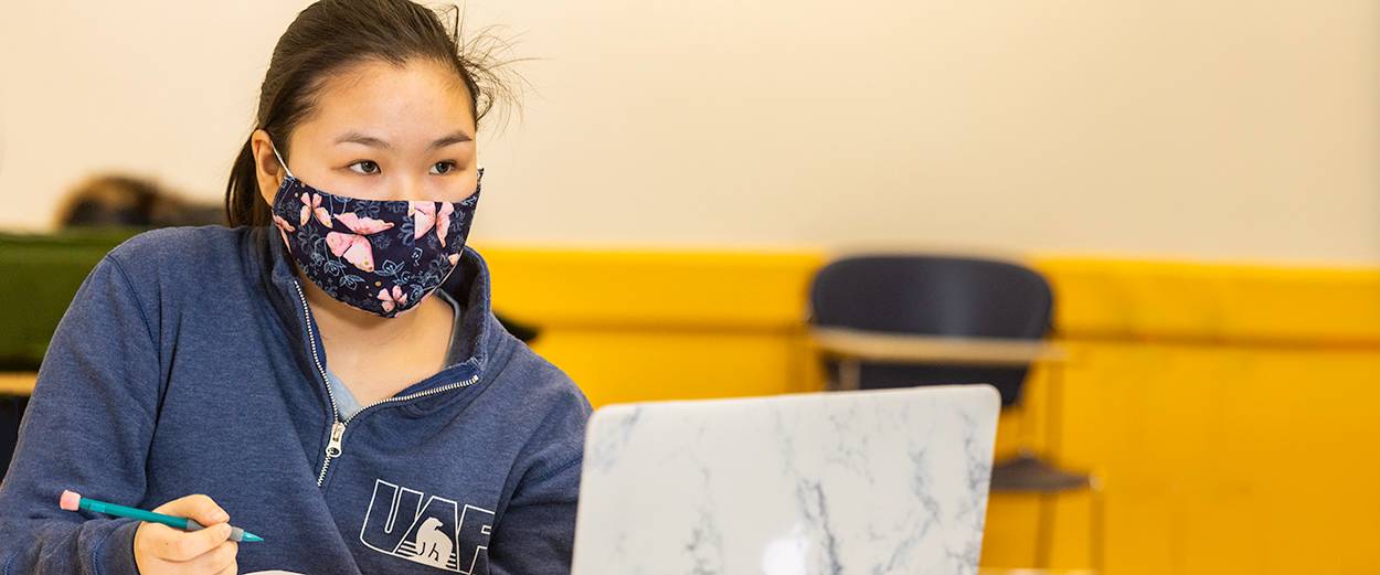 A student wearing a mask sits at a desk in a classroom with a laptop