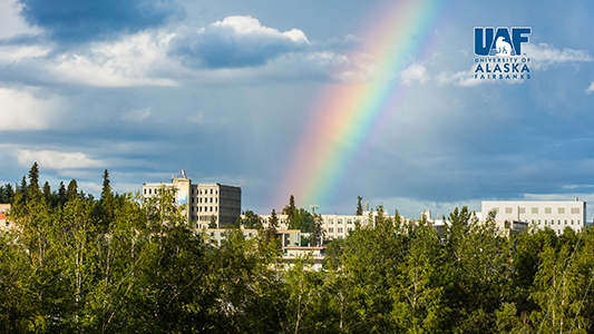 Looking east on the Fairbanks campus toward the Gruening Building on a summer day as a rainbow touches down in the background, with the UAF logo in the upper right corner.