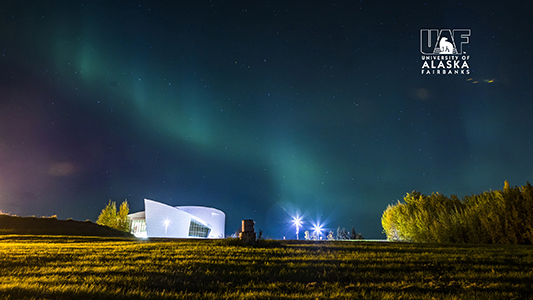 The aurora lights up the autumn sky over the UA Museum of the North, with the UAF logo in the upper right corner.
