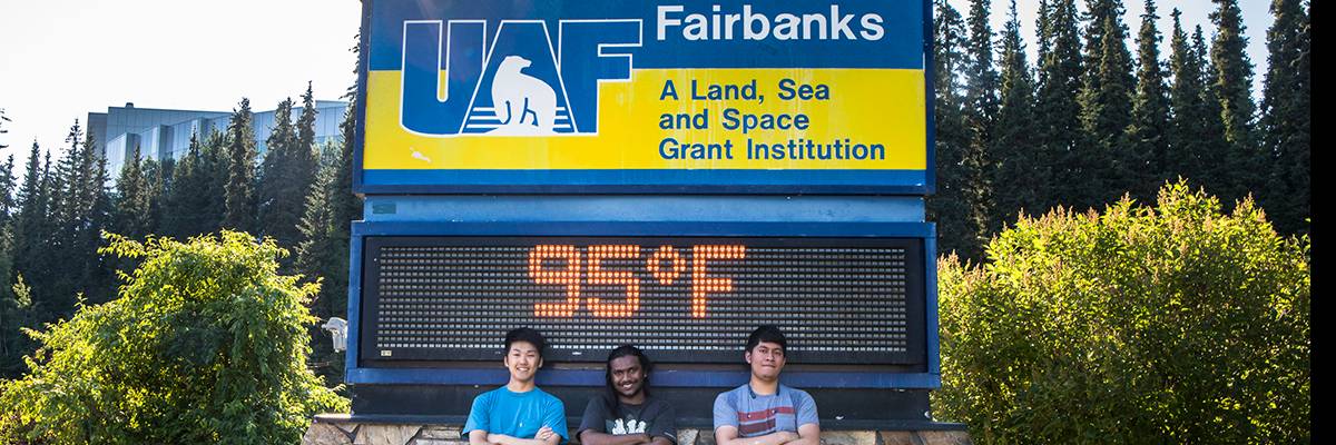 Three UAF students pose in front of the campus time and temperature sign, which reads 95 degrees, Fahrenheit